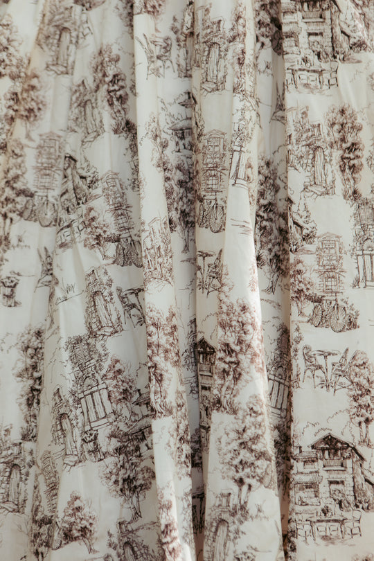 The Zaza Midi Dress in Brown Toile by Ivy City Co features cream-colored fabric with a brown toile pattern of trees, buildings, and nature, draping elegantly in soft folds.