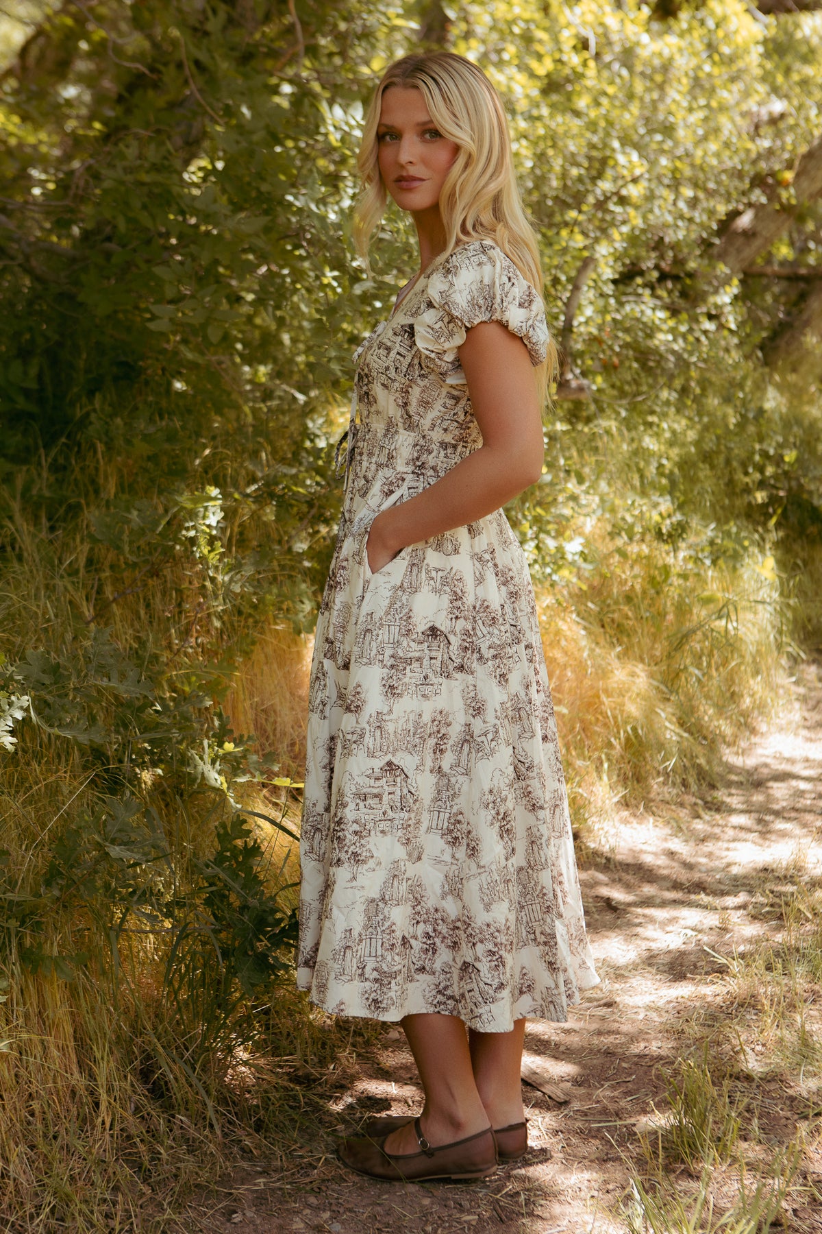 A woman with long blonde hair stands outdoors on a sunlit path, surrounded by greenery, wearing the Ivy City Co Zaza Midi Dress in Brown Toile with short sleeves and pockets, paired with brown sandals.