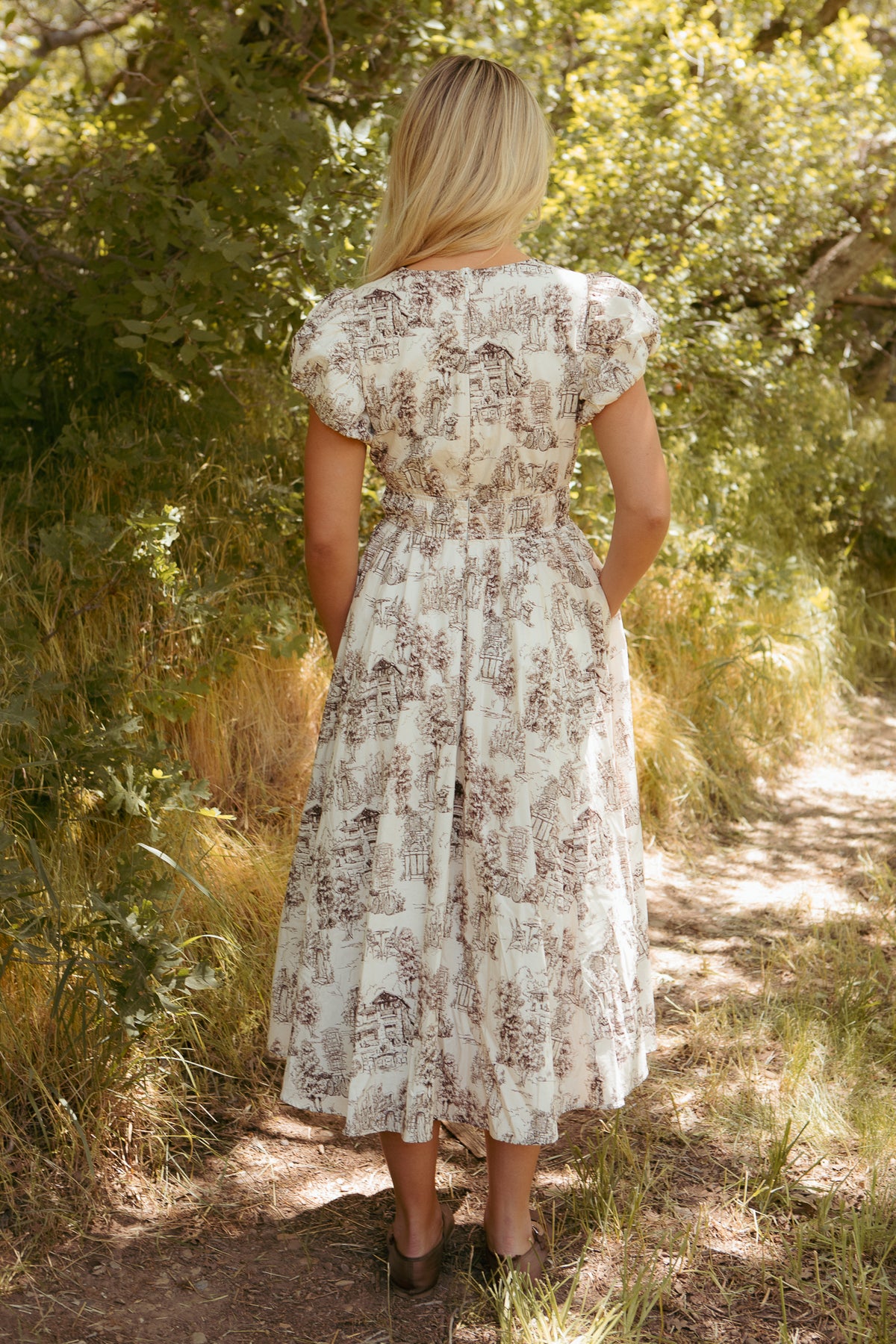 A blonde woman stands on a sunlit dirt path surrounded by greenery, wearing the Ivy City Co Zaza Midi Dress in Brown Toile, a white cotton midi dress with brown toile patterns.
