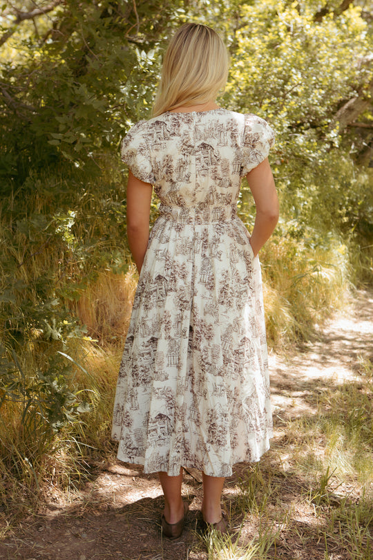 A blonde woman stands on a sunlit dirt path surrounded by greenery, wearing the Ivy City Co Zaza Midi Dress in Brown Toile, a white cotton midi dress with brown toile patterns.