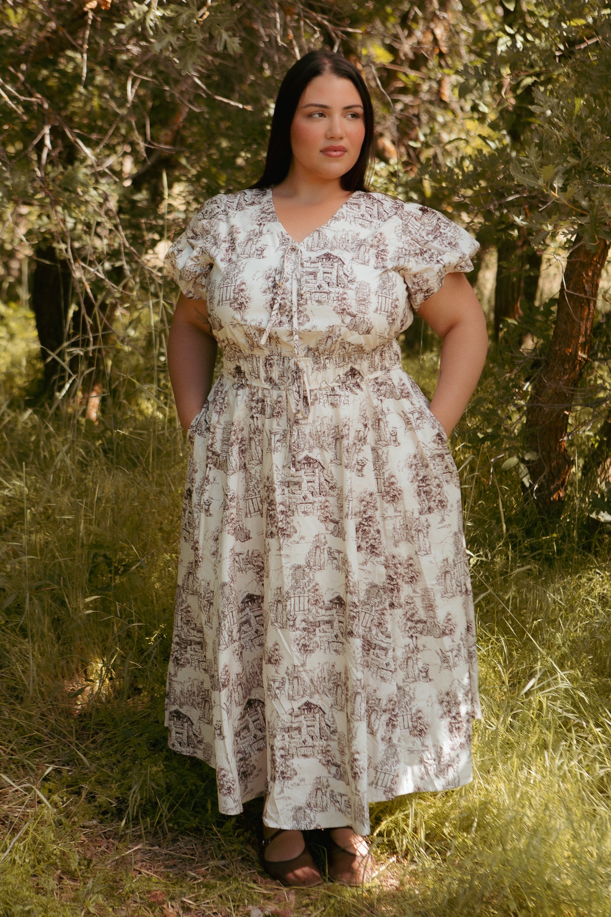 A woman with long dark hair stands in a sunlit forest, wearing the Ivy City Co Zaza Midi Dress in Brown Toile. The white cotton midi dress features brown scenic patterns. She gazes to the side, hands in her pockets amid grass and trees.