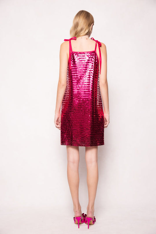 A woman is seen from behind, wearing the Ivy City Co Zelda Dress in Fuchsia Shingle Sequin—a shimmering, fuchsia sequin party dress featuring shoulder straps tied in bows—paired with matching pink high-heeled shoes. The background is plain and light-colored.