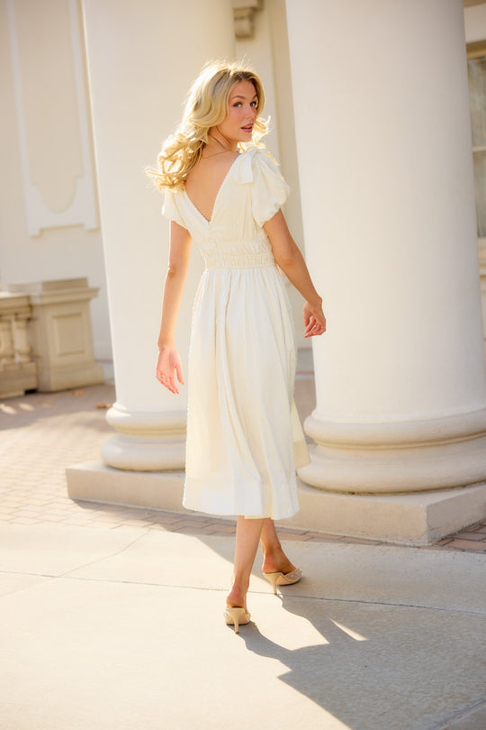 a woman in a white dress walking away and looking back outside in front of white pillars on cobblestone. 