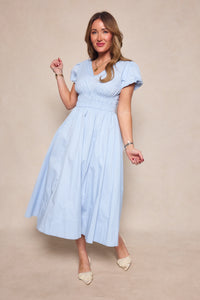 A woman poses against a neutral backdrop in the Ivy City Co Zella Dress in Sky Blue—a light blue, short-sleeve midi dress with a gathered waist and v-neckline—styled with bracelets, a necklace, and cream-colored shoes.