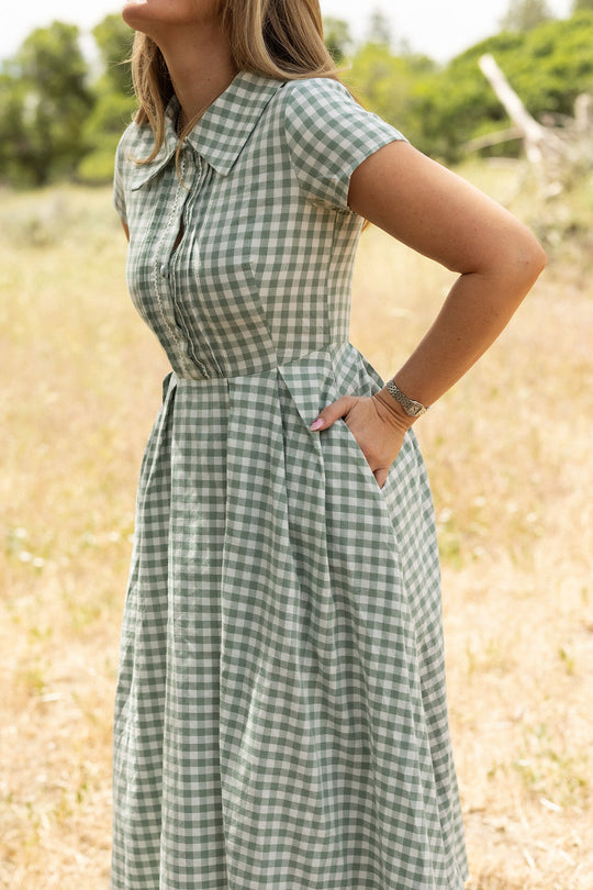 A woman stands outdoors on a grassy area with blurred trees in the background, hands in the pockets of her Ivy City Co Amber Dress in Sage Gingham—a cotton midi dress featuring a pleated skirt. Her face is not visible.