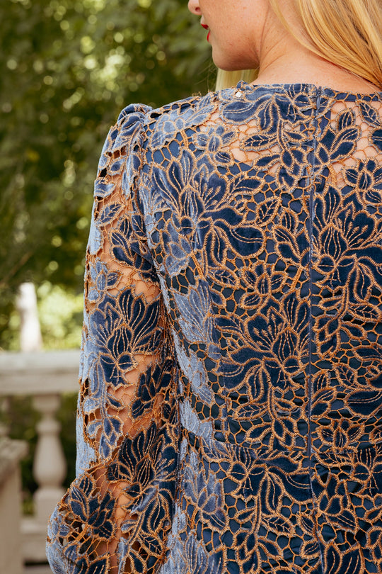 A close-up of a woman wearing the Janelle Dress in Golden Veil by Ivy City Co, adorned with intricate gold lace embroidery, standing outdoors beside a stone railing with lush greenery in the background.