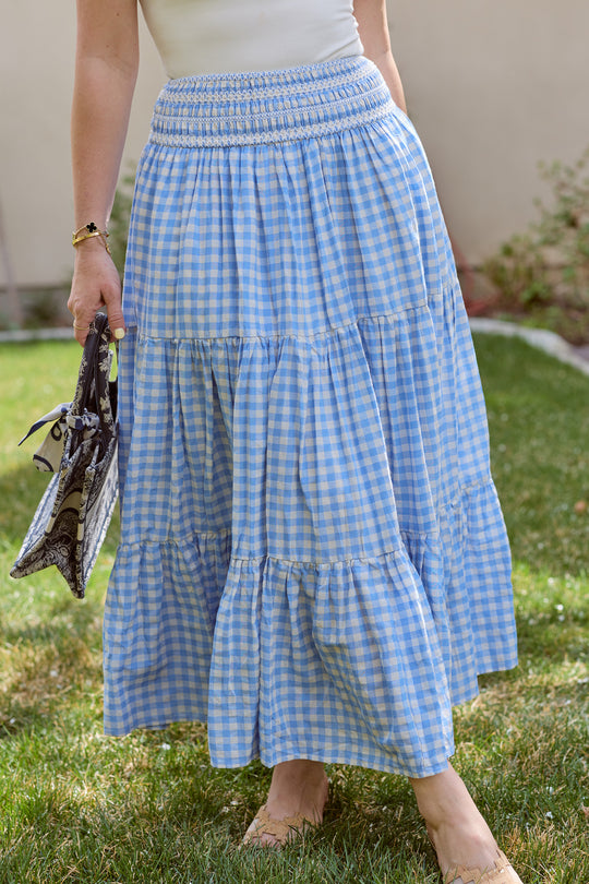 A woman’s lower body is visible as she stands on grass, holding a patterned bag and wearing the Zoey Skirt in Blue Gingham by Ivy City Co, which features a smocked waistband.