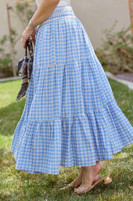 A person wearing the Ivy City Co Zoey Skirt in Blue Gingham with tan sandals stands on grass holding a patterned handbag, with a blurred backdrop of greenery and a beige wall.