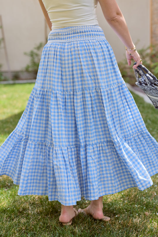 Someone stands on grass holding a patterned bag, wearing the Ivy City Co Zoey Skirt in Blue Gingham. Only the lower half of their body is visible.