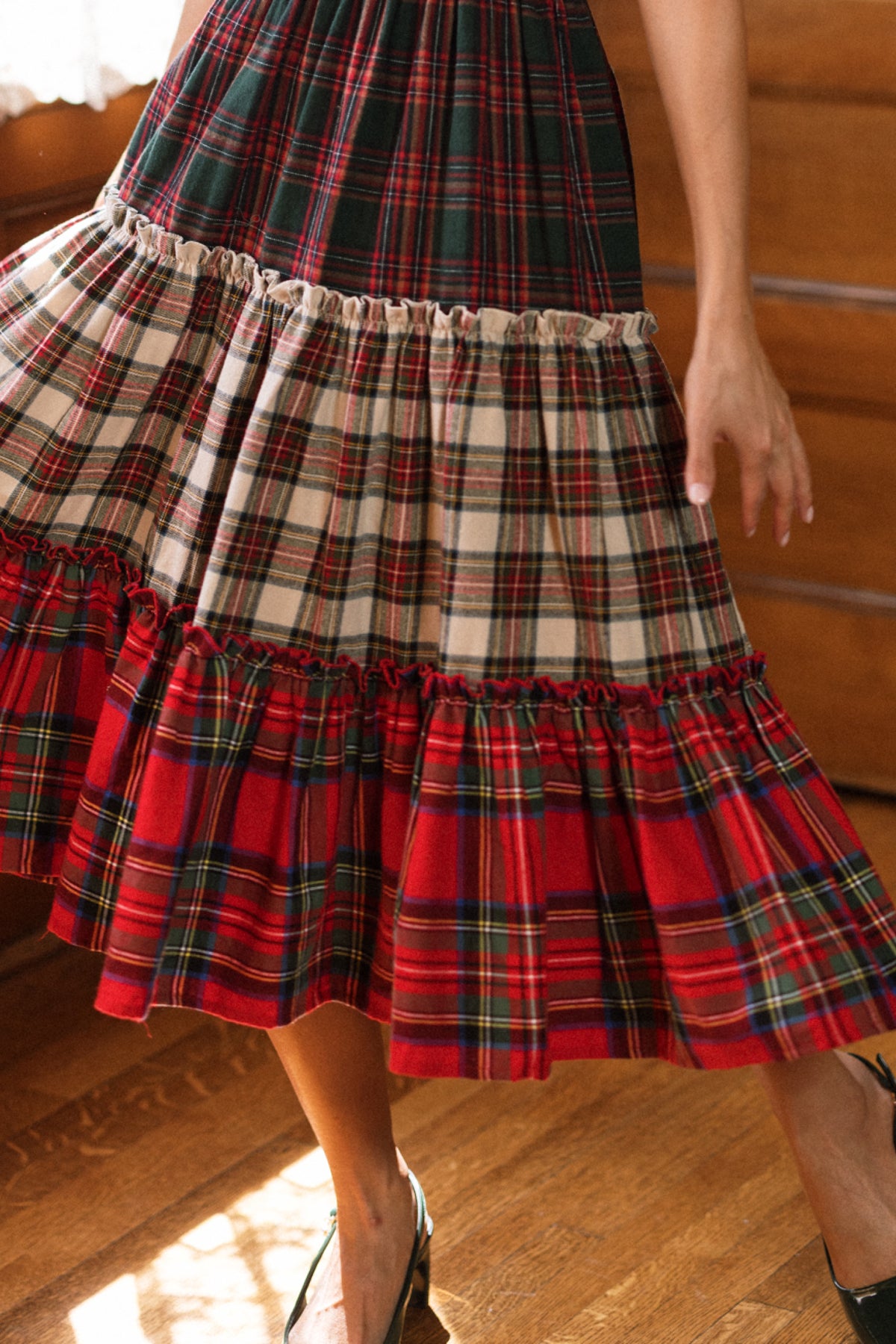 Wearing the Ivy City Co Zoey Skirt in Mixed Plaid, featuring green, cream, and red tiers, a person stands on a wooden floor with sunlight streaming in and one arm at their side. The cotton A-line skirt creates a soft silhouette that’s maternity friendly and ideal for holiday family gatherings.