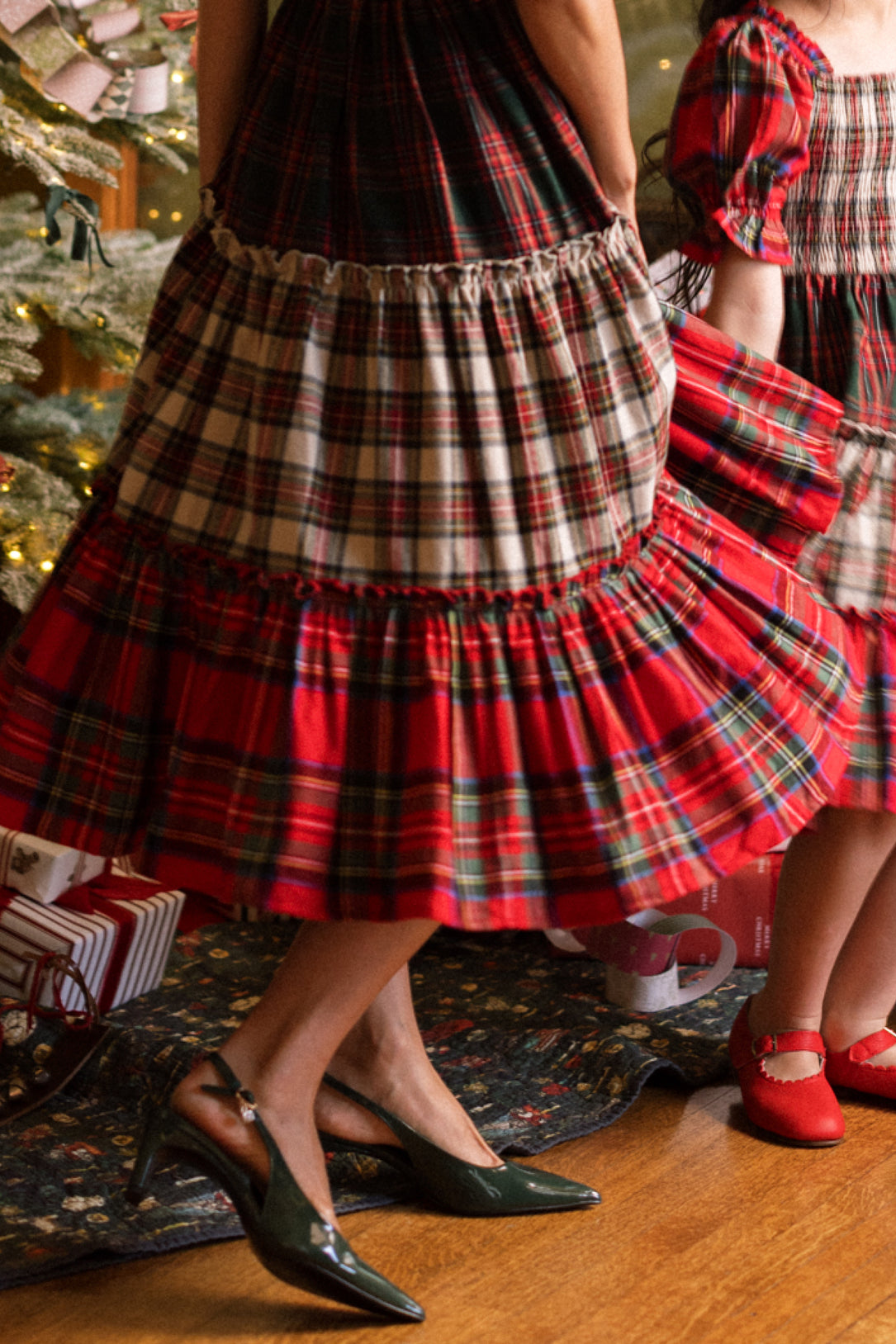 Two people, wearing the Ivy City Co Zoey Skirt in Mixed Plaid, stand near a decorated Christmas tree and wrapped gifts. One adult pairs the skirt with green heels, while a child wears red shoes. Only their lower halves are visible.