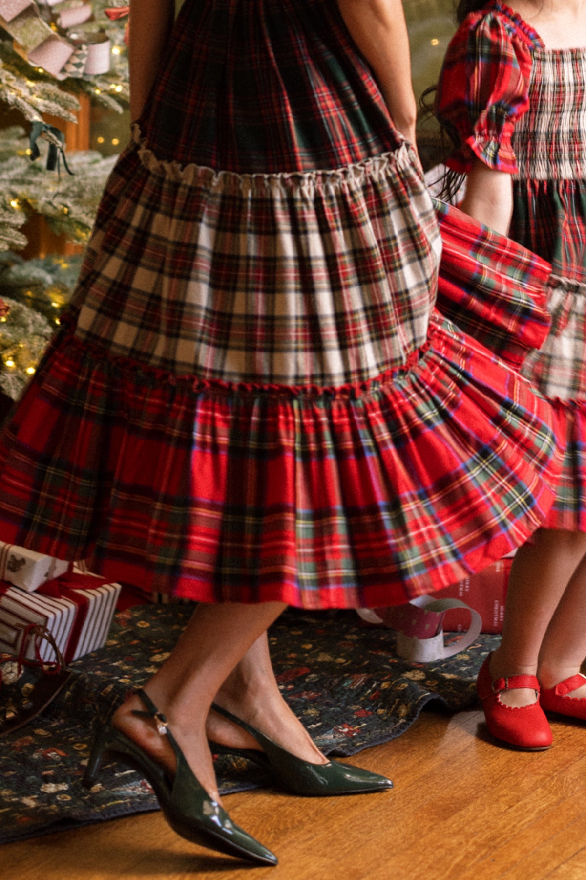 Two people, wearing the Ivy City Co Zoey Skirt in Mixed Plaid, stand near a decorated Christmas tree and wrapped gifts. One adult pairs the skirt with green heels, while a child wears red shoes. Only their lower halves are visible.