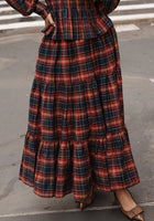 A person stands on a city sidewalk, their lower half visible in the Zoey Skirt in Twilight Plaid by Ivy City Co—a striking mix of red, navy, and yellow plaid styled with brown pointed-toe shoes for effortlessly chic fall fashion.