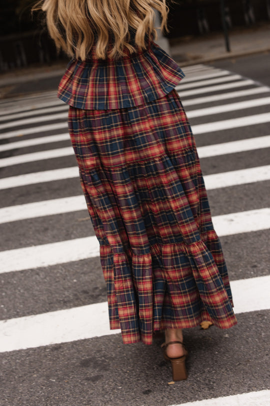 A woman walks across a crosswalk, seen from behind, wearing the Ivy City Co Zoey Skirt in Twilight Plaid—a tiered red and navy plaid maxi skirt with ruffle details, perfect for fall—paired with brown block-heel sandals and her blonde wavy hair flowing down.
