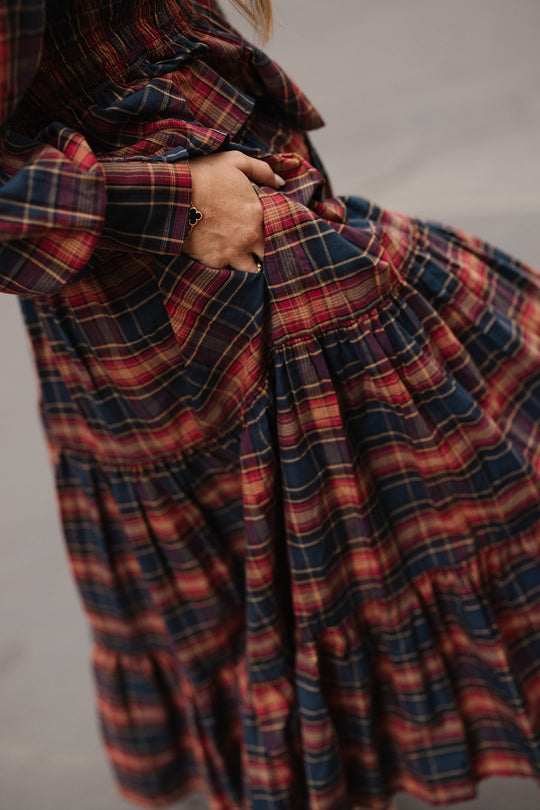 A person wearing the Zoey Skirt in Twilight Plaid by Ivy City Co holds a handful of the soft cotton A-line skirt, creating gentle folds in its red and navy plaid fabric. The neutral, out-of-focus background highlights an effortless fall fashion moment.