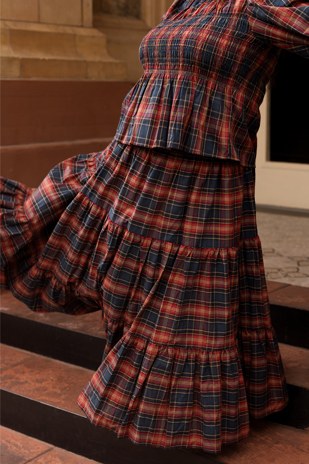 A person stands on brown steps wearing the Ivy City Co Zoey Skirt in Twilight Plaid—its striking red, navy, and yellow plaid pattern and ruffled details take center stage in the image, with the composition highlighting the skirt’s textured fabric and cozy fall fashion appeal while keeping the wearer’s face out of view.