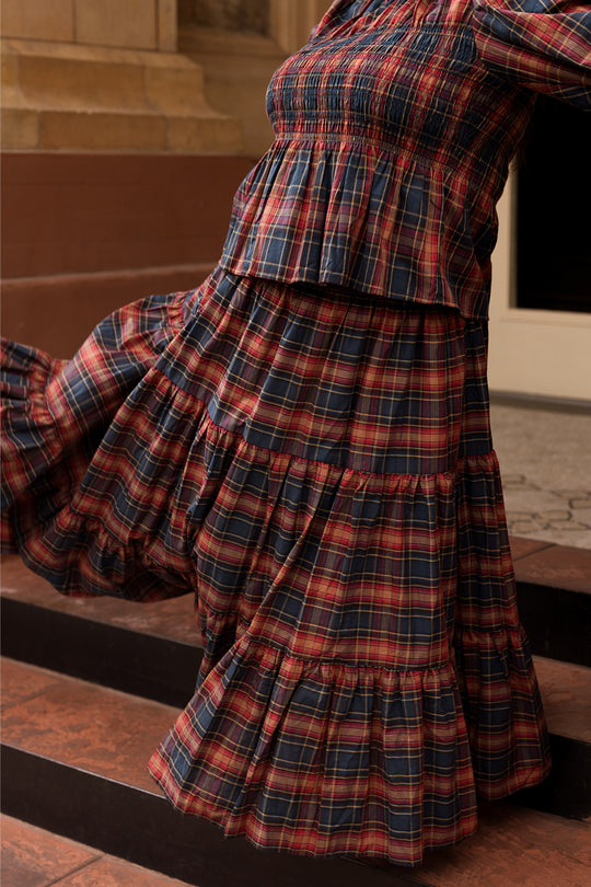 A person stands on brown steps wearing the Ivy City Co Zoey Skirt in Twilight Plaid—its striking red, navy, and yellow plaid pattern and ruffled details take center stage in the image, with the composition highlighting the skirt’s textured fabric and cozy fall fashion appeal while keeping the wearer’s face out of view.