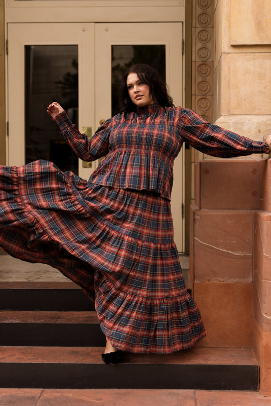 A woman poses dramatically on outdoor steps, showcasing the Ivy City Co Zoey Skirt in Twilight Plaid as it billows around her. Standing before tan stone walls and double doors, she captures the essence of classic fall fashion.