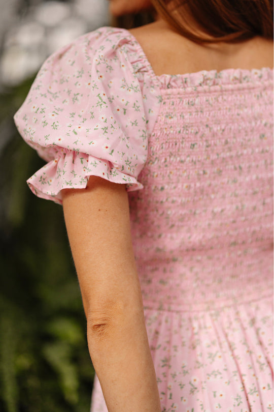 A woman is pictured from the back and side, standing outdoors in the Ivy City Co Valencia Dress in Pink Ditsy Floral. This lovely dress features puff sleeves, a smocked bodice, and a delicate feminine pattern, offering both charm and maternity-friendly comfort.