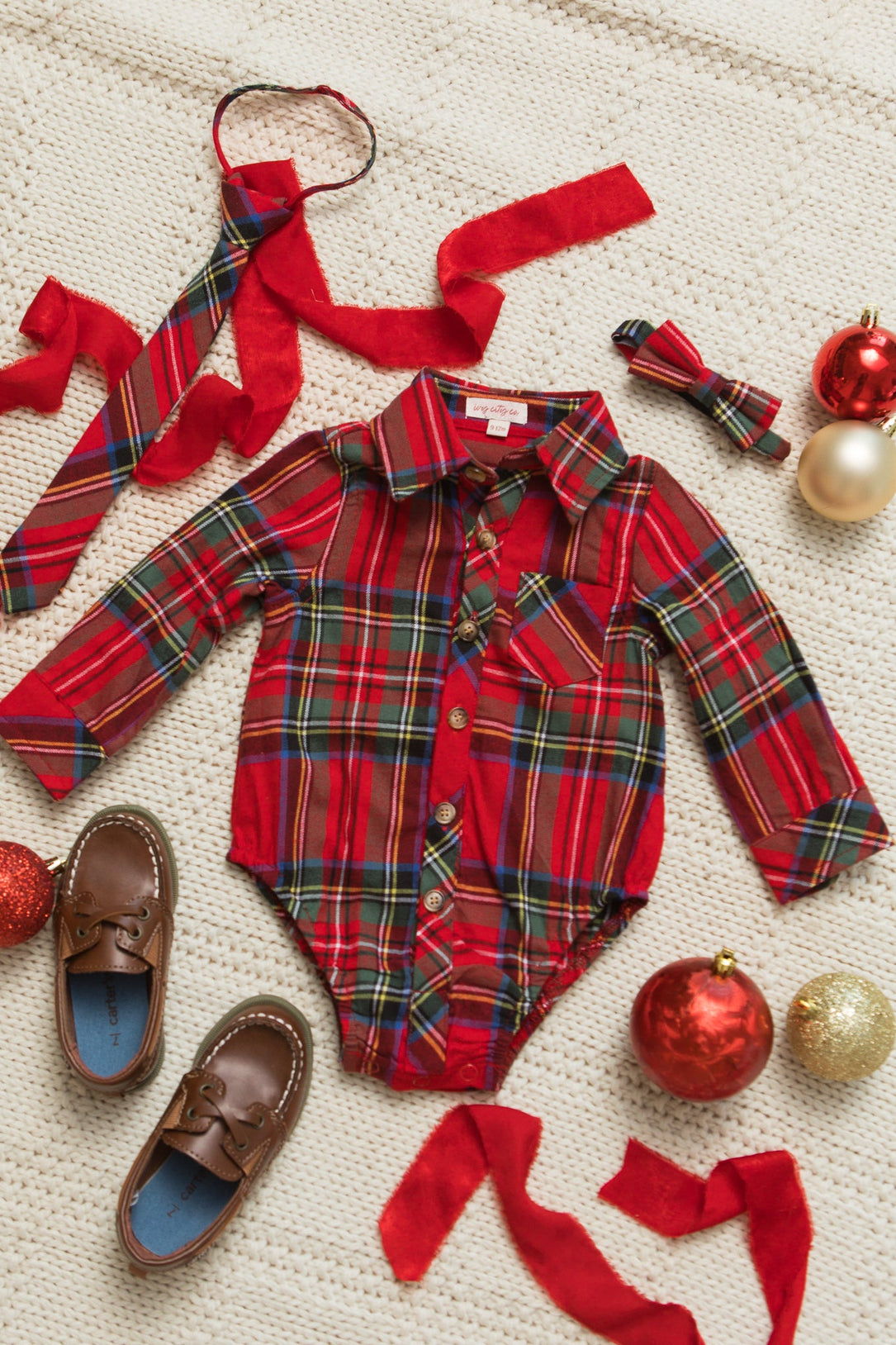 Flat lay featuring the Ivy City Co Baby Boys John Romper in Holiday Plaid, paired with brown baby loafers, a matching red cotton holiday plaid tie and bowtie, red ribbons, and Christmas ornaments arranged on a cream knit background.