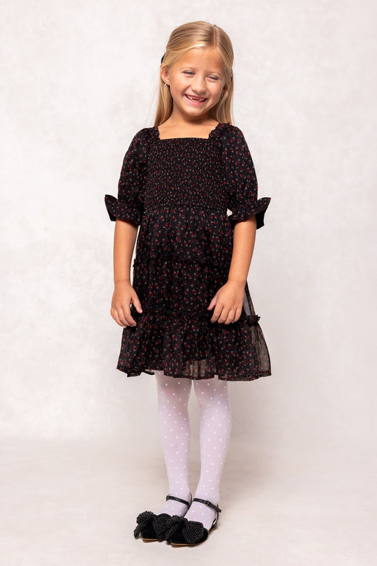 A smiling young girl with blonde hair styled half-up wears the Ivy City Co Mini Madeline Dress in Black Ditsy Floral, paired with white polka-dot tights and black shoes, standing against a light background.