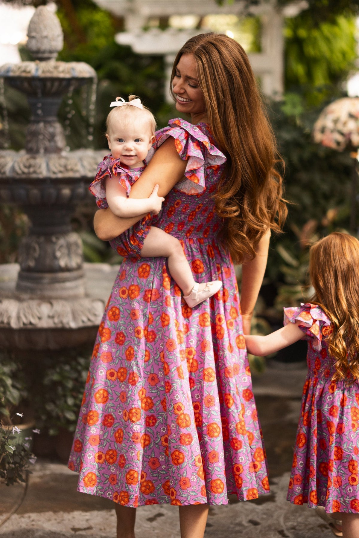 A woman holding a smiling baby—both dressed in matching purple and red dresses—stands beside a young girl in a similar dress near a stone fountain in a lush garden. The baby’s outfit, the Ivy City Co Baby Hattie Romper in Thistle Flower Poppy Woodblock, adds an extra touch of sweetness to this picturesque scene.