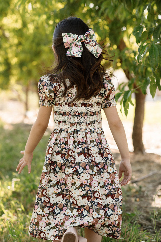 A young girl with long hair and a floral bow strolls outdoors among green trees, seen from behind on a sunny day. She wears the Ivy City Co Mini Sunday Best Dress in Midnight Garden, featuring a smocked bodice and puff sleeves in a matching floral print.