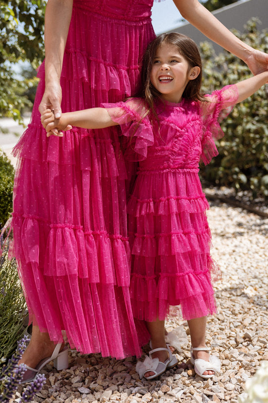 A smiling young girl in an Ivy City Co Mini Whimsical Dress in Fuchsia, featuring flutter sleeves and frilly details, holds hands with an adult wearing a matching dress as they stand together on a gravel path surrounded by greenery.