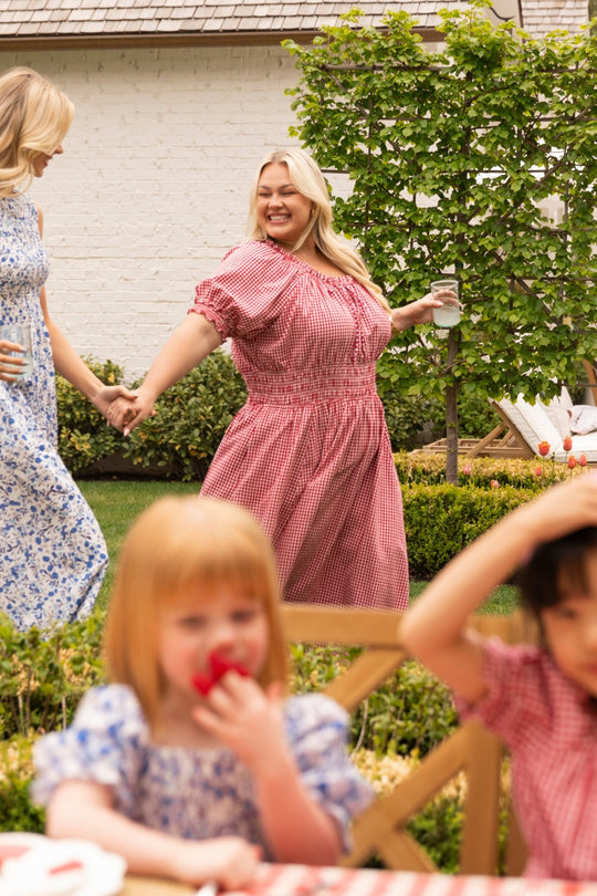 Two women holding hands and smiling in a garden—one wearing the Ivy City Co Jessie Dress in Red Gingham, which also serves as a comfortable maternity dress, and the other in a blue floral dress. In the foreground, two children sit at a table with one eating a strawberry.