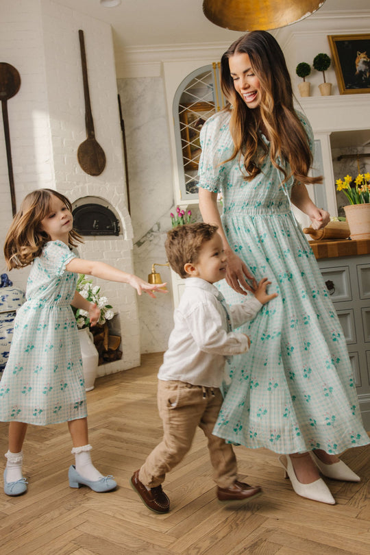 A woman wearing the Ivy City Co Jessie Dress in Bloom Gingham smiles in a kitchen with two young children. The little girl, also dressed in blue, and a boy in khakis playfully interact beside her on the wooden floor.
