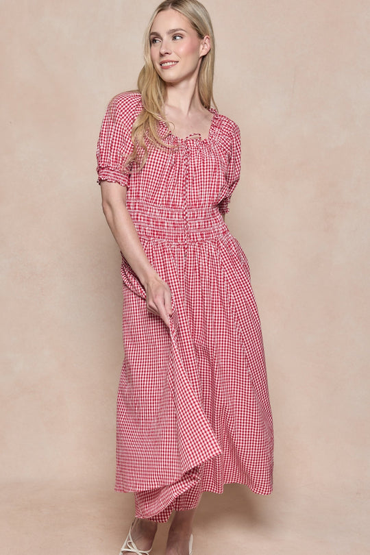 A woman wearing the Ivy City Co Jessie Dress in Red Gingham, featuring puff sleeves and nursing-friendly design, smiles as she looks to the side and holds one side of her skirt in front of a beige background. This stylish dress combines comfort and elegance for a charming look.