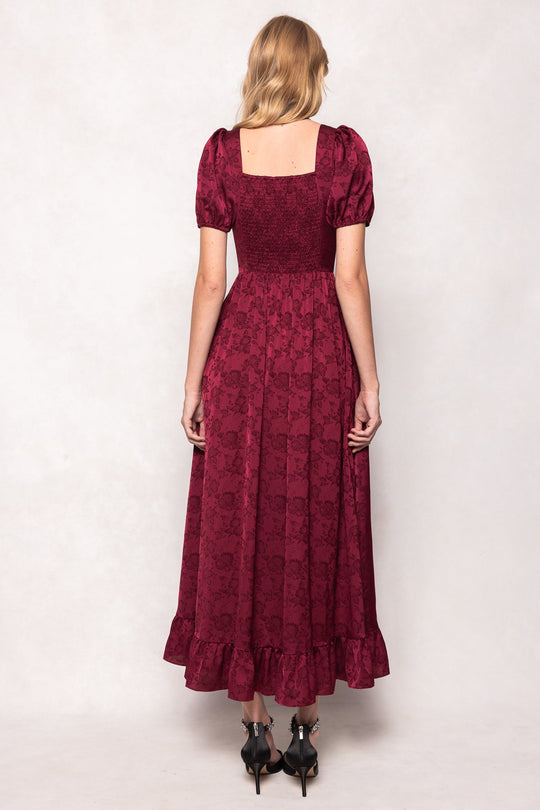 A woman with blonde hair stands facing away from the camera in the Amira Dress in Red by Ivy City Co, showcasing its short-sleeved, floral-patterned design with a ruffled hem, square neckline, and smocked back. She completes the look with black heeled shoes.