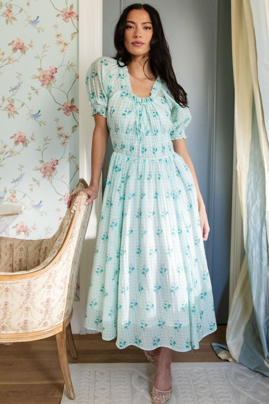 A woman stands indoors near a vintage-style chair and floral wallpaper, by a window with sheer curtains, wearing the Ivy City Co Jessie Dress in Bloom Gingham—a light blue, floral-patterned, puff-sleeve midi dress with a smocked waist. This maternity-friendly style is perfect for comfort and ease.