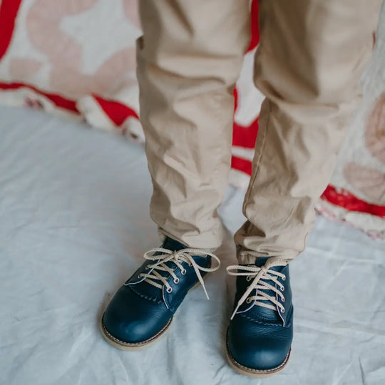 A child wearing beige pants and the Zimmerman Shoes Mini Boys Saddles in Navy, featuring classic leather construction and a stylish fringe tongue, stands on a white surface with a red and beige patterned blanket in the background—perfectly dressed for formal events.