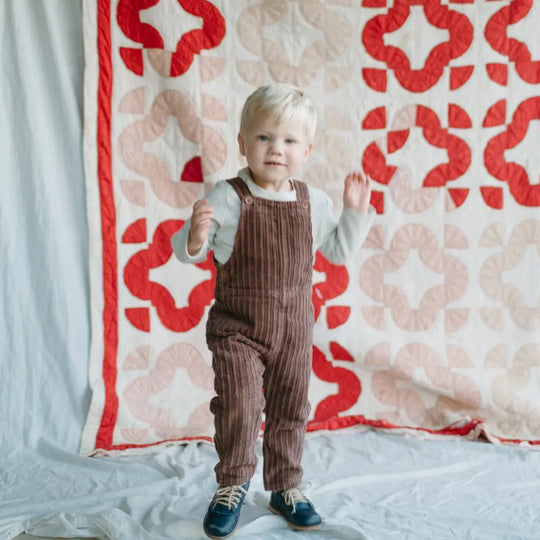A young child with light hair, dressed in brown overalls and wearing Zimmerman Shoes’ Mini Boys Saddles in Navy—with their classic leather design and fringe tongue—stands on a light fabric before a red and white patterned quilt backdrop.