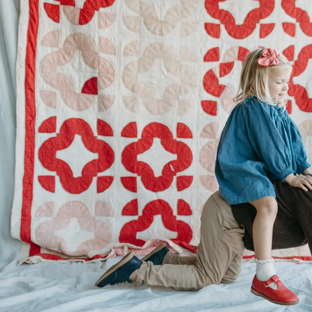 A young girl in a blue dress and red shoes, wearing Zimmerman Shoes’ Mini Boys Saddles in Navy—a classic navy leather saddle shoe with a fringe tongue—sits on a person’s back, who is kneeling on all fours in front of a red and white quilted backdrop.