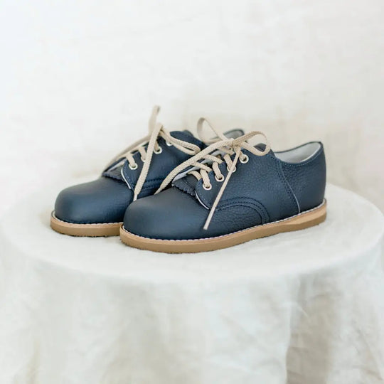 A pair of Zimmerman Shoes Mini Boys Saddles in Navy, featuring small, dark blue lace-up uppers and tan soles reminiscent of classic saddle shoes, are displayed on a round, white draped surface against a light background.