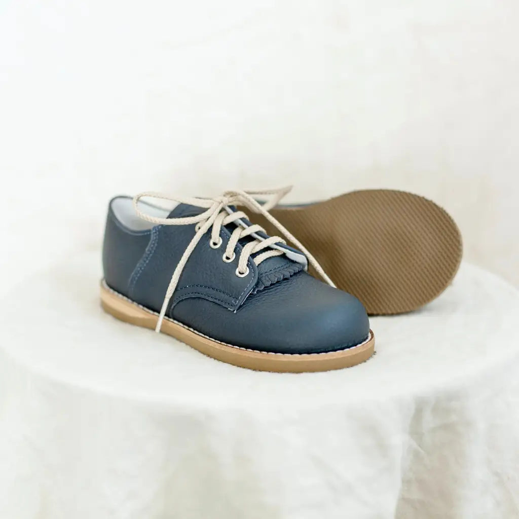 The Mini Boys Saddles in Navy by Zimmerman Shoes, featuring navy leather with a fringe tongue and tan soles, are shown on a white cloth surface—one shoe upright, the other on its side—making them ideal for formal events.