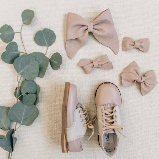 A pair of Zimmerman Shoes’ Mini Boys Saddles in Sand & Fog, four beige hair bows in assorted sizes, and a sprig of green eucalyptus leaves are artfully arranged on a light fabric background.