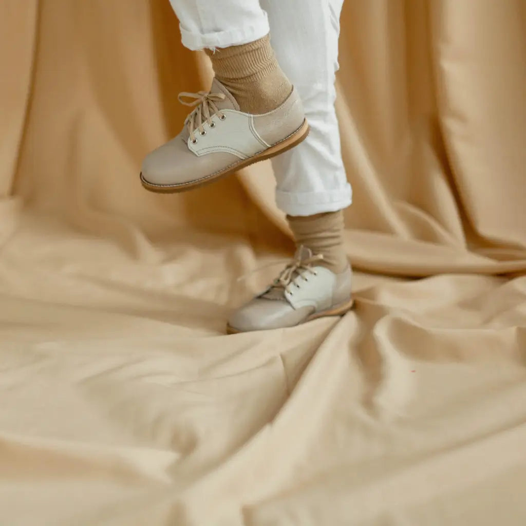 A person wearing Zimmerman Shoes’ Mini Boys Saddles in Sand & Fog steps onto a soft, beige fabric backdrop. Only the lower legs and feet are visible, and the outfit blends seamlessly with the neutral-toned setting.