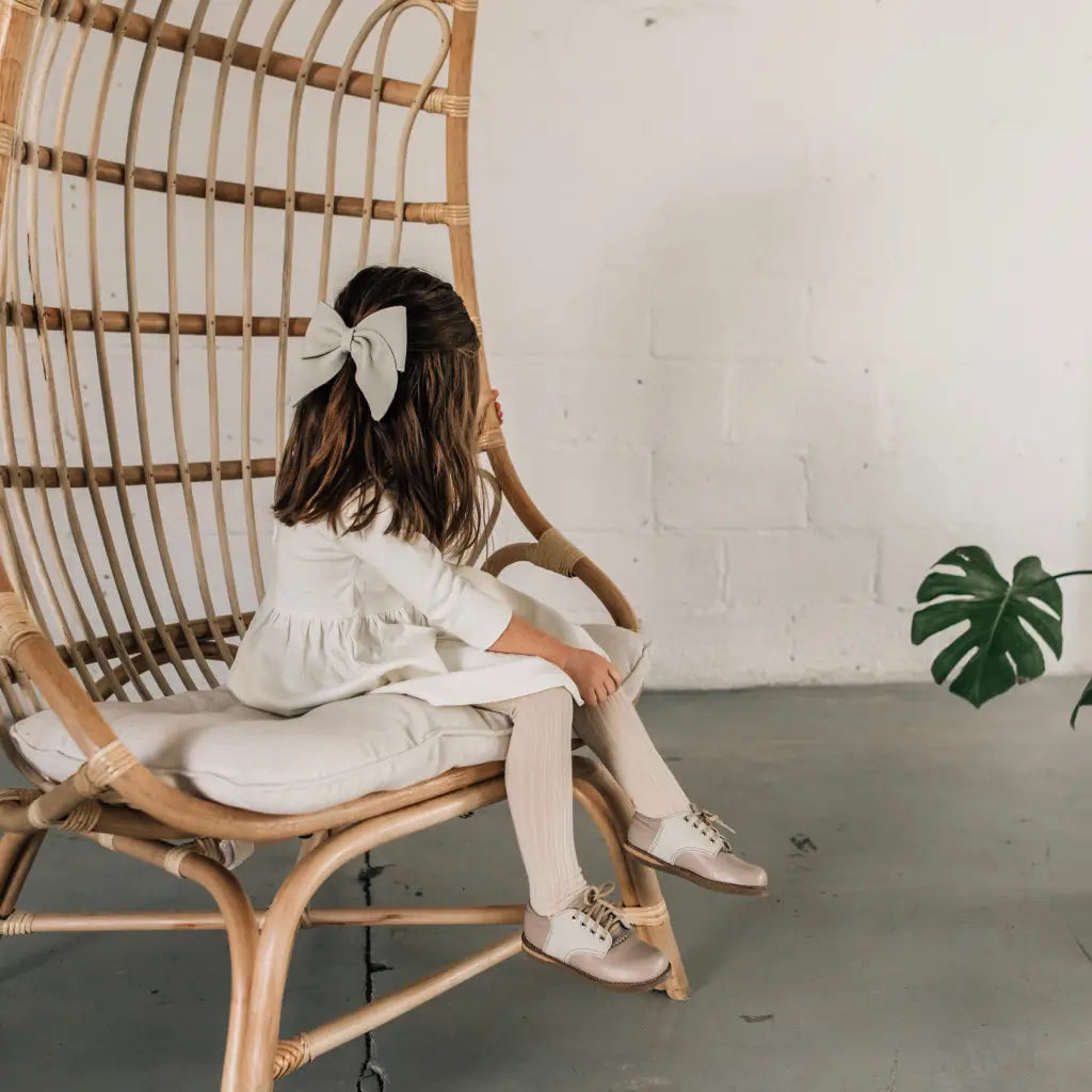 A young girl with a large bow in her hair sits on a rattan chair, facing away. She wears a white dress, tights, and Zimmerman Shoes Mini Boys Saddles in Sand & Fog. A green Monstera plant rests on the floor beside her. The background is minimal and light.