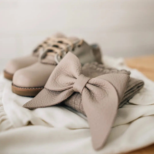 Mini Boys Saddles in Sand & Fog by Zimmerman Shoes rest in the background, complemented by a matching leather bow and folded fabric in the foreground, all arranged elegantly on a soft white cloth.