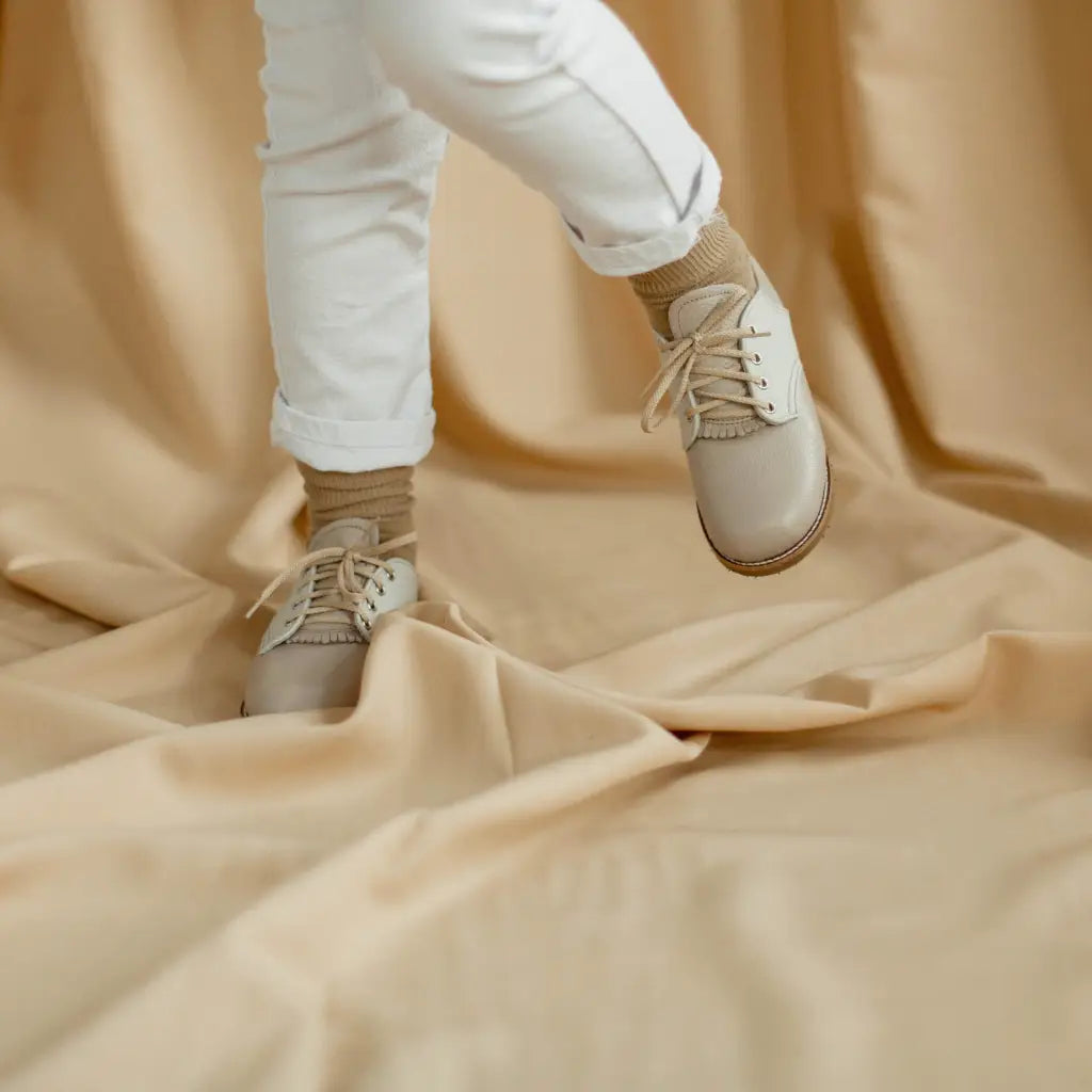 A child wearing white pants and beige socks stands on crumpled beige fabric, lifting one foot off the ground to showcase Zimmerman Shoes’ Mini Boys Saddles in Sand & Fog.