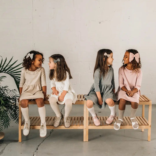 Four young girls sit on wooden benches against a white wall, wearing pastel dresses and bows in their hair as they smile and chat together. Each pair of formal shoes—one featuring the Zimmerman Shoes Mini Boys Saddles in Sand & Fog, and another styled with beige leather flats—adds a charming touch to their delightful outfits. A green plant is visible on the left side of the image.