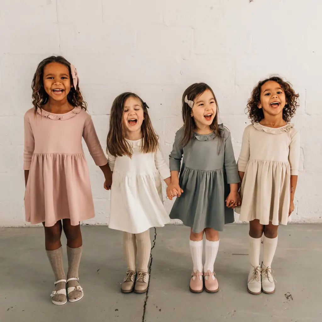 Four young girls stand in a row, smiling and holding hands. They wear long-sleeved dresses in pink, white, gray, and beige, each paired with matching socks and Zimmerman Shoes’ Mini Boys Saddles in Sand & Fog formal shoes, set against a plain white wall.