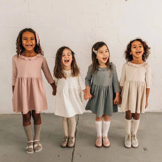 Four young girls stand in a row, smiling and holding hands. They wear long-sleeved dresses in pink, white, gray, and beige, each paired with matching socks and Zimmerman Shoes’ Mini Boys Saddles in Sand & Fog formal shoes, set against a plain white wall.