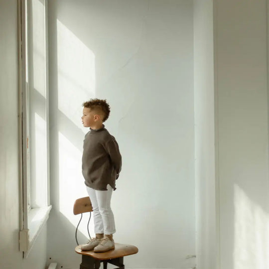 A young child wearing Zimmerman Shoes’ Mini Boys Saddles in Sand & Fog stands on a chair, gazing out of a sunlit window in a minimalist room with white walls and soft natural light.