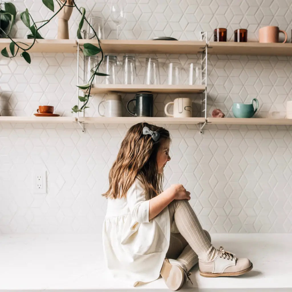 A young girl with long hair and a bow, wearing Zimmerman Shoes’ Mini Boys Saddles in Sand & Fog, sits on a white counter facing left. Behind her are shelves with mugs, cups, and a vine plant against a white, patterned backsplash.
