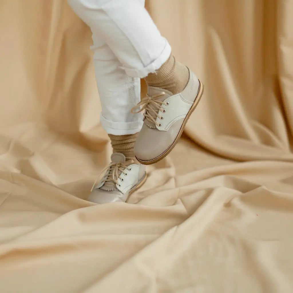 A person models the Zimmerman Shoes Mini Boys Saddles in Sand & Fog, paired with white pants and beige socks, standing on a beige fabric backdrop with one leg slightly raised.