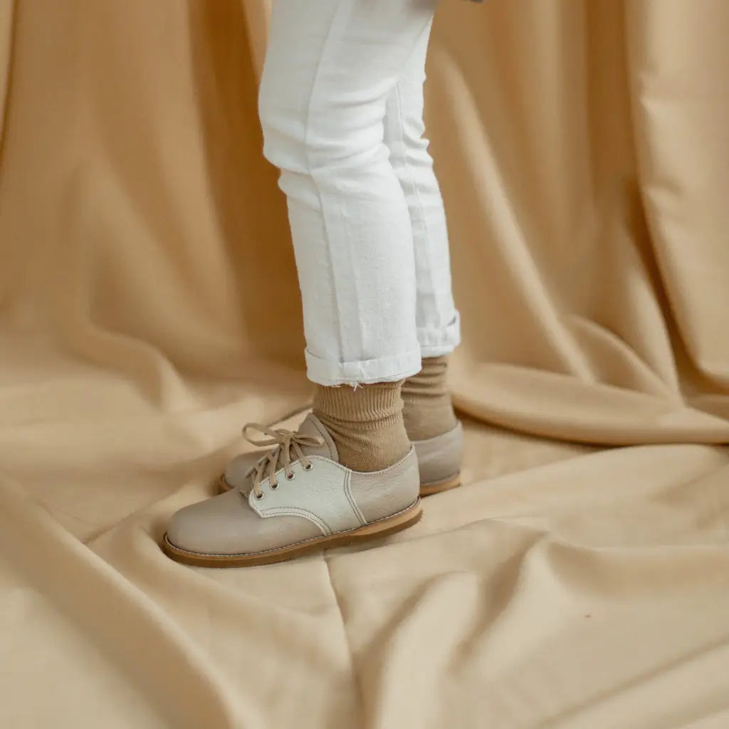 A child wearing white pants and tan socks models the Zimmerman Shoes Mini Boys Saddles in Sand & Fog, standing on a beige draped fabric background. Only the lower legs and feet are visible.